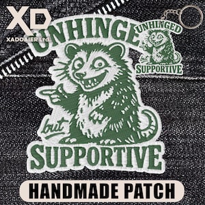 May include: Embroidered green patch with a raccoon design and the words "UNHINGED but SUPPORTIVE." A small keychain with a matching raccoon design is also visible. The patch is labeled "HANDMADE PATCH."