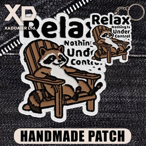 May include: Embroidered patch and keychain featuring a raccoon relaxing in a wooden chair. The patch reads "Relax, Nothing Is Under Control." The keychain has a similar design. The patch has a white border and the text "HANDMADE PATCH".