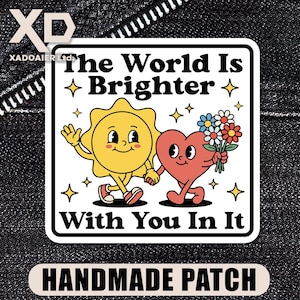 May include: A handmade patch with a white border featuring a cartoon sun and heart holding hands. The text "The World Is Brighter With You In It" is above and below the characters. The patch also has the text "HANDMADE PATCH".