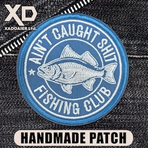 May include: A round, blue embroidered patch with the text "AIN'T CAUGHT SHIT FISHING CLUB" encircling a detailed fish illustration. The patch also features the text "HANDMADE PATCH".