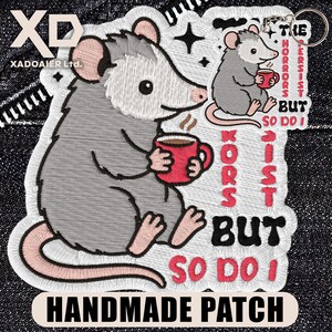 May include: Embroidered patch featuring a gray opossum holding a red mug with coffee. The patch includes the text "The horrors persist but so do I" and "Handmade Patch".