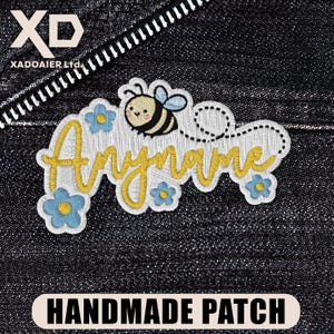 May include: Embroidered patch with the text "Anyname" in yellow script, a cartoon bee, and blue flowers. The patch is outlined in white and reads "HANDMADE PATCH" at the bottom. The background is dark denim.