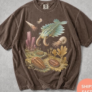 Peut inclure: Un t-shirt à manches courtes couleur espresso orné d'une impression graphique détaillée de vie marine préhistorique, incluant des trilobites, des ammonites et un grand arthropode, sur un paysage sous-marin rocheux et texturé avec des coraux.