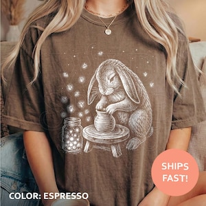 May include: An espresso-colored t-shirt featuring a white line-art graphic of a rabbit sitting at a pottery wheel, shaping a clay pot. Beside the rabbit is a glass jar filled with glowing fireflies, surrounded by small sparkling stars.