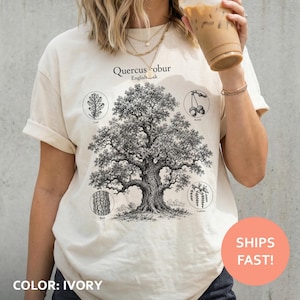 Peut inclure: Un t-shirt ivoire arborant une illustration botanique noire d'un Quercus robur, ou chêne pédonculé. Le motif comprend des croquis détaillés d'une feuille, de glands, d'écorce et de chatons, avec le texte Quercus robur English oak imprimé au-dessus.