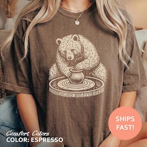 May include: A dark brown short-sleeve t-shirt featuring a cream-colored graphic print of a bear sitting at a pottery wheel and shaping a clay pot. The design is centered on the chest area of the garment, which has a relaxed, loose-fitting silhouette.