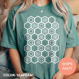 May include: A teal short-sleeve t-shirt featuring a white graphic print of a repeating honeycomb pattern. The design consists of interconnected hexagons, with some containing a six-pointed star motif. The shirt has a crew neckline.