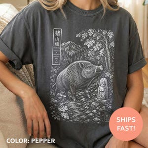 May include: Pepper-colored t-shirt featuring a detailed black and white illustration of a wild boar, a mountain scene, and Japanese text. The graphic includes a stone statue and a path. The shirt has short sleeves.