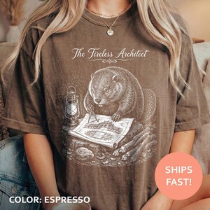 May include: An espresso-coloured short-sleeve t-shirt featuring a white graphic of a beaver working at a desk with a lantern and architectural blueprints. The text above the illustration reads The Tireless Architect in a cursive white font.
