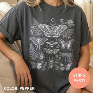 May include: Pepper-coloured t-shirt with a white illustration of insects, butterflies, and plants. The design includes a large moth, a bee, and several butterflies, along with ferns and other botanical elements.