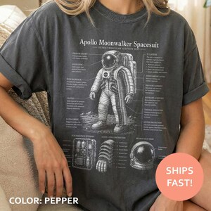 May include: A charcoal gray t-shirt featuring a white graphic print of an Apollo Moonwalker Spacesuit. The design includes a central astronaut illustration with technical labels, a backpack, a glove, and a helmet, all detailed with text.