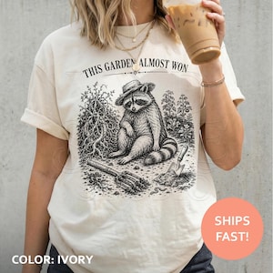 May include: An ivory t-shirt featuring a black ink illustration of a raccoon wearing a hat, sitting in a garden with a shovel and a rake. The text above the raccoon reads, THIS GARDEN ALMOST WON. The design includes detailed plants and soil.