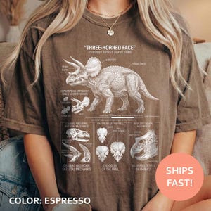 May include: Espresso-colored t-shirt featuring a detailed illustration of a Triceratops, labeled "THREE-HORNED FACE." The design includes anatomical diagrams and text. The shirt's color is noted as "ESPRESSO."