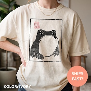 May include: An ivory short-sleeve t-shirt featuring a black ink-style graphic of a frog inside a rectangular border. Above the frog is a red square stamp with Asian characters. The design has a textured, hand-printed appearance on the fabric.