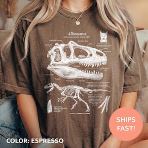 May include: A brown short-sleeve t-shirt featuring a white graphic print of an Allosaurus skull and skeleton. The design includes scientific labels and text detailing the Allosaurus fragilis, Marsh, 1877, with anatomical diagrams of the dinosaur.