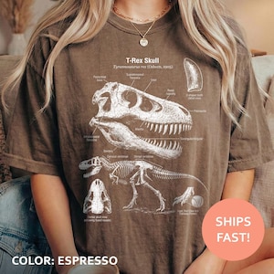 May include: Espresso-colored t-shirt featuring a detailed white illustration of a T-Rex skull and skeleton with labeled anatomical features. The text "T-Rex Skull Tyrannosaurus rex (Osborn, 1905)" is printed above the graphic.