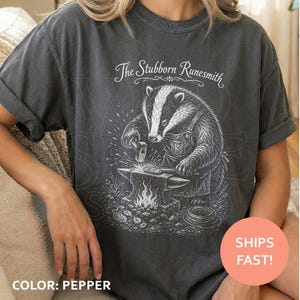May include: A charcoal gray short-sleeve t-shirt featuring a white graphic illustration of a badger blacksmith working at an anvil. The text above the badger reads The Stubborn Runesmith. The design includes sparks and metalworking tools.