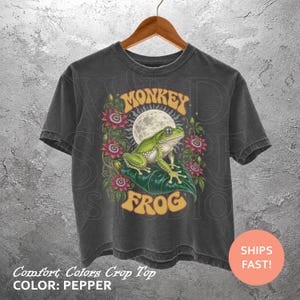May include: A charcoal gray short-sleeve t-shirt featuring a graphic print of a green frog sitting on a leaf under a full moon, surrounded by purple and red passion flowers. The text MONKEY FROG is printed in yellow above and below the illustration.