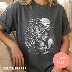 May include: A charcoal gray short-sleeve t-shirt featuring a white graphic illustration of an owl perched on a pottery wheel, shaping a clay pot. The scene includes a full moon, clouds, and a lit candle, rendered in a detailed, sketch-like style.