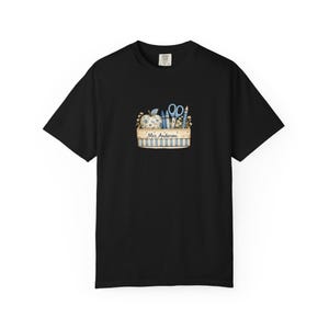 May include: A black short-sleeve t-shirt featuring a central graphic of a rectangular crate with blue and white stripes. The crate contains a floral apple, blue crayons, scissors, and a pencil, with the text Mrs. Anderson printed on the front.