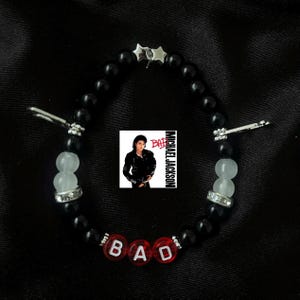 Op de afbeelding: Zwart kralenarmband met witte en heldere kralen, zilveren accenten en het woord "BAD" in rode letters. Een kleine afbeelding van Michael Jackson is ook inbegrepen. De armband heeft een stervormige bedel.