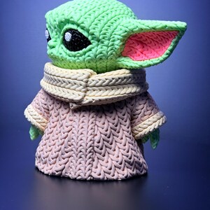 K&ouml;nnte beinhalten: Eine gr&uuml;n-rosa Figur eines Charakters mit gro&szlig;en Ohren und schwarzen Augen, die einen hellrosa Pullover und einen cremefarbenen Schal tr&auml;gt. Die Figur ist eine stilisierte Darstellung eines beliebten Charakters aus einem Science-Fiction-Franchise.