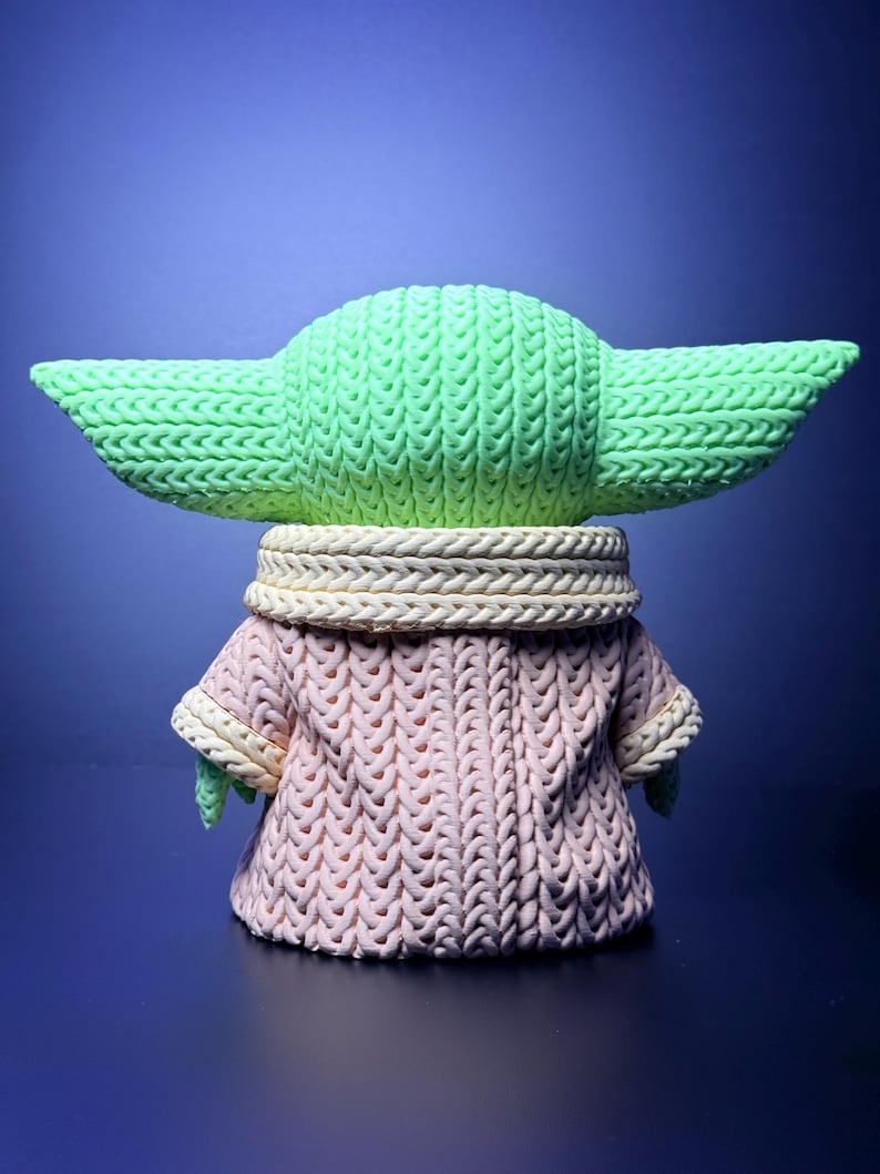 K&ouml;nnte beinhalten: Eine gr&uuml;ne Figur eines Charakters mit gro&szlig;en Ohren, die einen rosa Strickmantel mit cremefarbenem Kragen tr&auml;gt. Die Figur steht vor einem blauen Hintergrund und zeigt die detaillierte Textur des Strickdesigns.