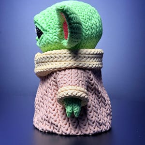 K&ouml;nnte beinhalten: Eine gr&uuml;ne, gestrickte Figur eines Charakters mit gro&szlig;en Ohren und einem rosa gef&uuml;tterten Ohrinneren. Die Figur tr&auml;gt einen rosa Pullover mit cremefarbenem Kragen und Manschetten. Die Figur ist eine stilisierte Darstellung eines beliebten Charakters.