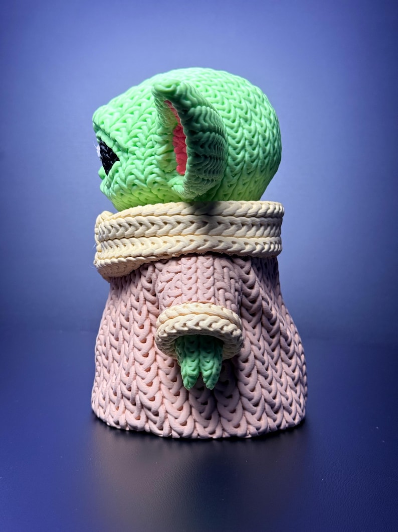 K&ouml;nnte beinhalten: Eine gr&uuml;ne, gestrickte Figur eines Charakters mit gro&szlig;en Ohren und einem rosa gef&uuml;tterten Ohrinneren. Die Figur tr&auml;gt einen rosa Pullover mit cremefarbenem Kragen und Manschetten. Die Figur ist eine stilisierte Darstellung eines beliebten Charakters.