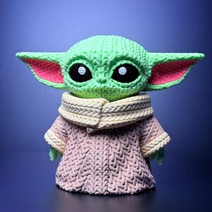 K&ouml;nnte beinhalten: Eine gr&uuml;ne Figur eines Charakters mit gro&szlig;en Ohren und schwarzen Augen, die ein rosa-cremefarbenes Strickoutfit tr&auml;gt. Die Figur hat ein strukturiertes Aussehen mit sichtbaren Strickmustern auf Kopf und Kleidung. Der Hintergrund ist ein Blauton.