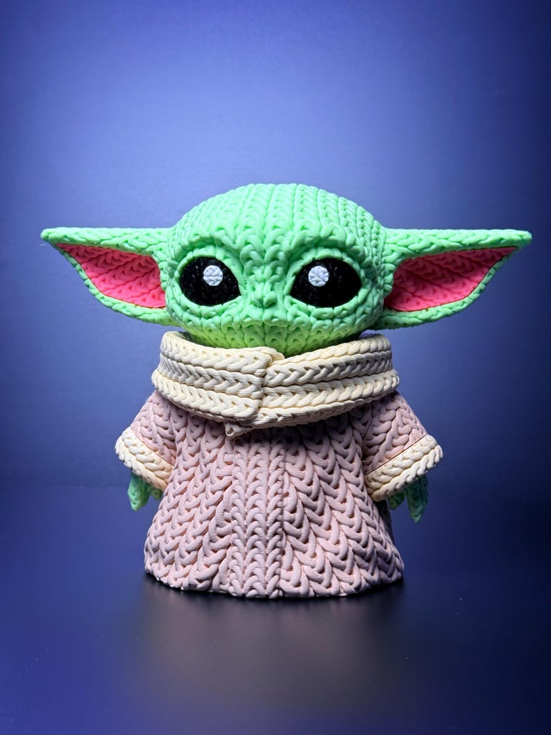 K&ouml;nnte beinhalten: Eine gr&uuml;ne Figur eines Charakters mit gro&szlig;en Ohren und schwarzen Augen, die ein rosa-cremefarbenes Strickoutfit tr&auml;gt. Die Figur hat ein strukturiertes Aussehen mit sichtbaren Strickmustern auf Kopf und Kleidung. Der Hintergrund ist ein Blauton.