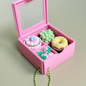 May include: A pink plastic box with a hinged lid and a clear window, containing four miniature food-shaped erasers: a white cupcake with blue accents, a cluster of light green grapes, a yellow donut with sprinkles, and three pink strawberries.