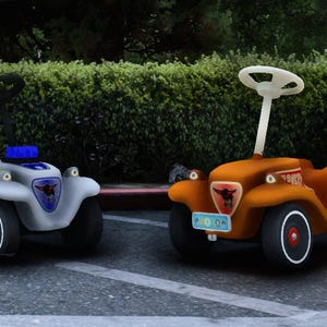 May include: Three colorful toy cars with trailers are parked on a paved surface. The cars are in shades of orange, light gray, and purple. Each car has a steering wheel and a trailer. The wheels have red accents. The background features green foliage.