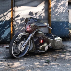 Puede incluir: Motocicleta personalizada con una combinación de colores gris y negro. Cuenta con una gran rueda delantera con muchos radios, un carenado y alforjas. La motocicleta está estacionada sobre una superficie de hormigón frente a un garaje.