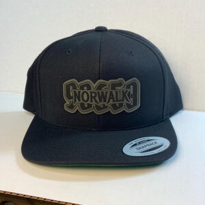 May include: A black snapback cap featuring a dark olive green patch on the front with the text NORWALK in a stylized font. The hat has a flat brim with a circular silver and black sticker that reads THE AUTHENTIC SNAPBACK YUPOONG SINCE 1974.