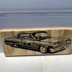 May include: A rectangular light-coloured wooden plaque featuring a detailed black laser-engraved illustration of a classic two-door coupe car. The wood grain is visible throughout the surface, providing a natural texture behind the car graphic.