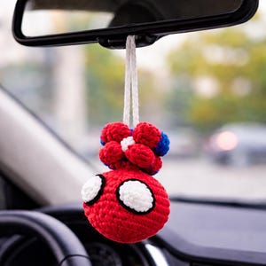 Peut inclure: Un porte-bonheur de voiture crocheté rouge, blanc et bleu, suspendu au rétroviseur. Le charme présente un corps rond rouge avec des accents blancs en forme d'yeux et un groupe de pompons rouges et bleus.