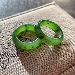 May include: Two jade rings with a vibrant green hue and dark speckles. The rings are smooth and polished, resting on a textured, tan surface. The rings are a classic band style, perfect for everyday wear.
