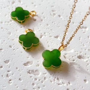 May include: A gold-toned necklace with a four-leaf clover pendant, alongside two matching clover charms. The clover pendants are a vibrant green with gold-colored trim, set against a textured white surface.