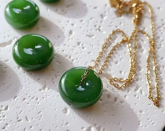 May include: A gold chain necklace with a round, polished, deep green jade pendant. Several loose, similar green jade stones are scattered on a textured white surface. The necklace is presented in a well-lit setting, showcasing the jade's colour.