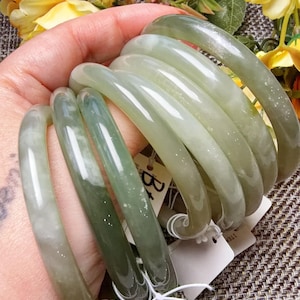 May include: A stack of five smooth, polished, light green jade-colored bangles with varying degrees of translucency and subtle white inclusions. Each bangle features a rounded profile and is attached to a small white paper tag with black text.