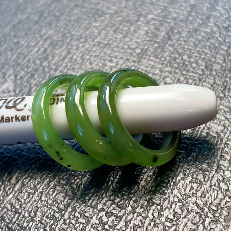 May include: Three translucent green jade rings. The rings are stacked on a white marker. The rings have small black inclusions. The rings are a light green color.