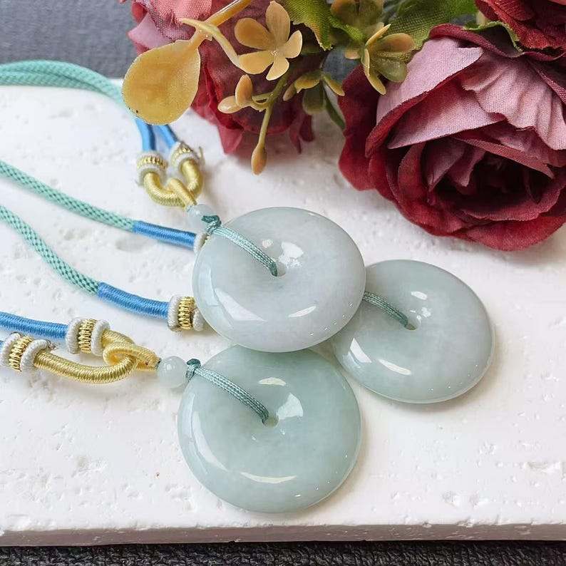 May include: Three jade donut pendants with light green cords. The pendants are a pale green colour and have a smooth, polished surface. The cords are blue and green, with gold and white accents. The background includes red and pink flowers.
