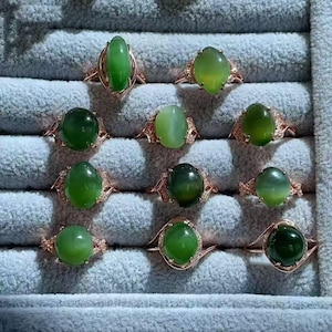 May include: A collection of ten rings featuring smooth, oval-shaped green gemstones set in rose-colored metal bands. Each ring displays a unique shade of green, ranging from light lime to deep forest tones, arranged in a grid pattern.