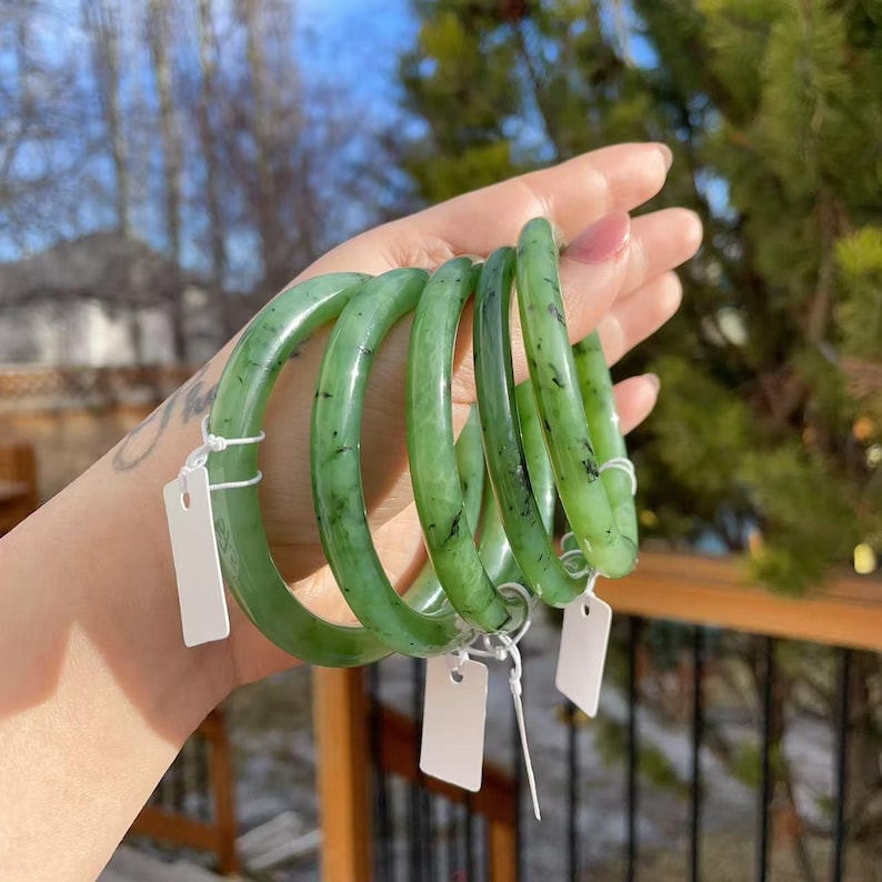 May include: Several jade green bangles are displayed. The bracelets are a vibrant green color with dark flecks. Each bangle is round and smooth, with a polished finish. White tags are attached to the bracelets.