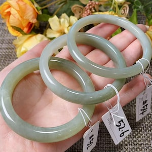 May include: Three smooth, pale green jade bangles with rounded profiles. Each bangle features a small white paper tag attached with a thin white string, displaying handwritten numbers and text. The bangles have a polished, translucent finish.