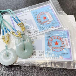 May include: Jade pendant necklaces with light green circular pendants. The pendants are strung on blue and gold cords. Two certificates are visible in the background, with Chinese text and red stamps.