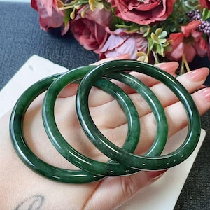 Puede incluir: Tres brazaletes de jade verde oscuro. Las pulseras lisas y pulidas se exhiben sobre un fondo de flores rosas y rojas. Los brazaletes son redondos y tienen un acabado brillante, que refleja la luz.