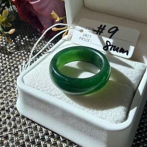 May include: A solid, smooth, dark green jade band ring with a polished finish. The ring is presented in a white jewelry box with a white tag attached by a string. The tag features the printed text ITEM #9 and the handwritten text 8rum.