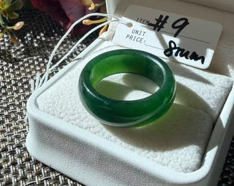 May include: A solid, smooth, dark green jade band ring with a polished finish. The ring is presented in a white jewelry box with a white tag attached by a string. The tag features the printed text ITEM #9 and the handwritten text 8rum.